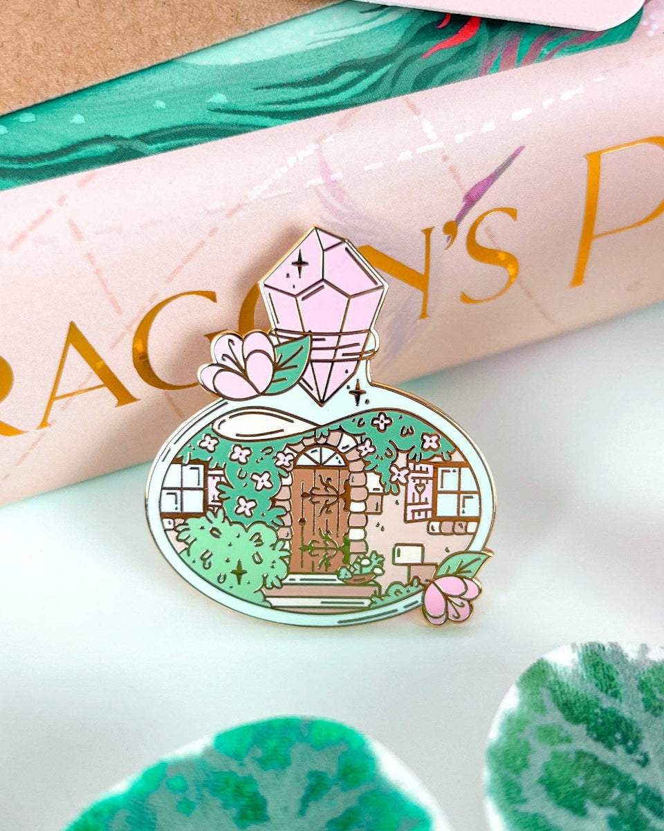 A cottage door with plants, vines, windows, and stairs inside a potion bottle.  The potion bottle is decorated with flowers and closed with a pink gem.  The design is on a hard enamel pin with gold plating.  The pin is leaning backwards onto the spine of a pink and gold book.