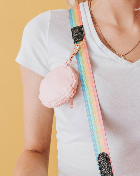 A pastel pink 3.45" (inches) circular treat pouch attached to a sassy stripes pastel rainbow with black details and gold-toned clasps worn by platinum blonde model with a v-neck white t-shirt.