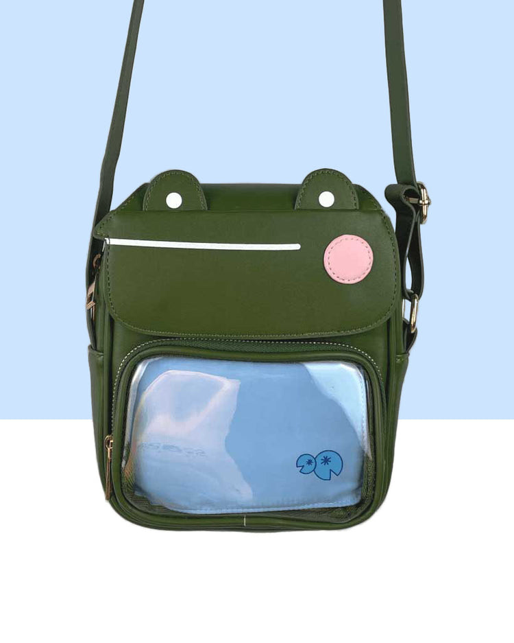 An olive green frog mini crossbody purse featuring son the frog.  The top flap is the face of a frog that opens to the inside.  The frog has a pink cheek and a straight line mouth.  The front pocket is clear with a blue insert.  The insert has two lily pads on it.  This also doubles as an ita bag.