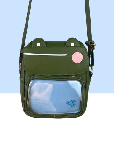 An olive green frog mini crossbody purse featuring son the frog.  The top flap is the face of a frog that opens to the inside.  The frog has a pink cheek and a straight line mouth.  The front pocket is clear with a blue insert.  The insert has two lily pads on it.  This also doubles as an ita bag.