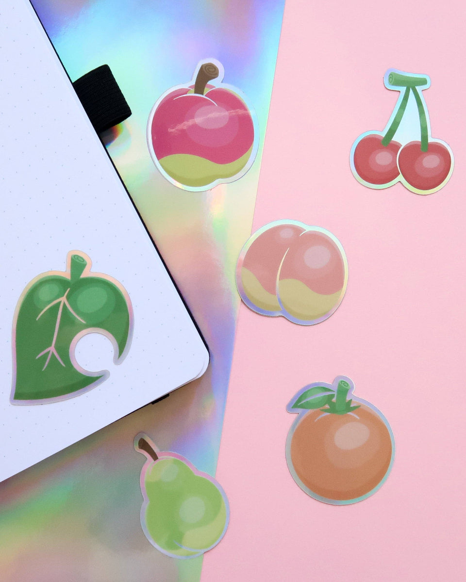 A group of holographic vinyl stickers shaped like animal crossing new horizons fruit including apple, cherry, peach, pear, orange, and the furniture leave.