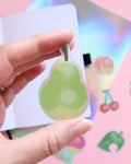A holographic vinyl sticker shaped like an animal crossing new horizons pear held between two fingers.  In the background there is a peach sticker, cherry, apple, and leaf stickers sitting on a pink and holographic background.  