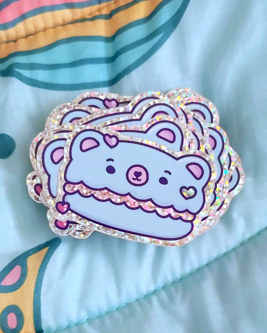 A blue and pink macaron cookie digital artwork shaped like a kawaii bear with hot pink, glitter green, and pastel pink heart sprinkle candies decorating it.  The macaron filling is pink glitter.  All of this is printed on a glossy vinyl sticker.  This photo shows a big pile of the sticker on top of a light blue blanket.