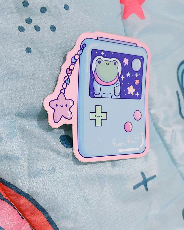 A kawaii matte mirror with orange chrome border vinyl sticker featuring a blue game boy style gaming handheld console with pink buttons and an unhappy green cross hair button. The image also has a happy pink star charm with three hearts. The screen features a frog astronaut in space with stars and moon. An original Prism Pear Designs design. Shown at an angle