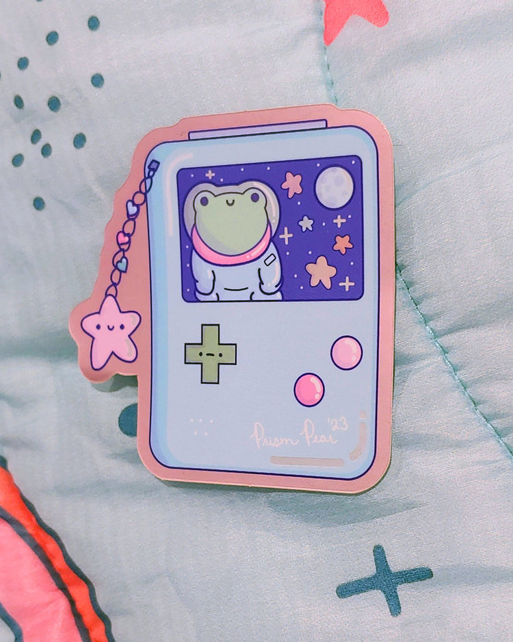 A kawaii matte mirror with orange chrome border vinyl sticker featuring a blue game boy style gaming handheld console with pink buttons and an unhappy green cross hair button. The image also has a happy pink star charm with three hearts. The screen features a frog astronaut in space with stars and moon. An original Prism Pear Designs design. Shown at an angle.