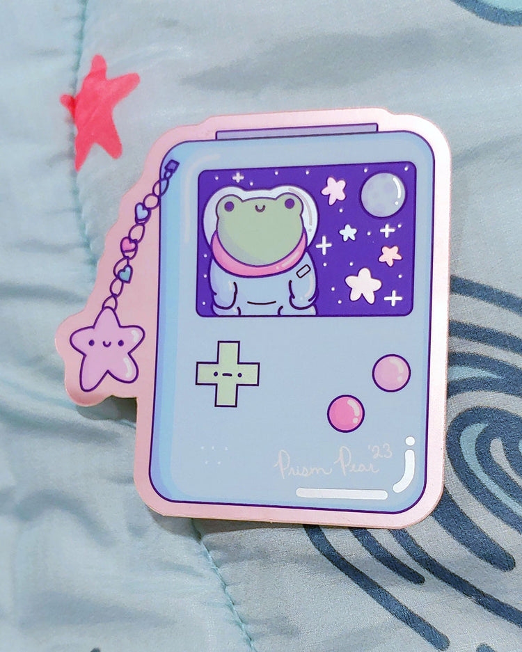 A kawaii matte mirror with orange chrome border vinyl sticker featuring a blue game boy style gaming handheld console with pink buttons and an unhappy green cross hair button. The image also has a happy pink star charm with three hearts. The screen features a frog astronaut in space with stars and moon. Displayed at an angle to chow mirror affect.