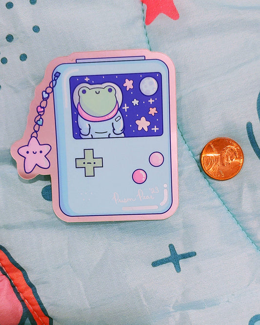 A kawaii matte mirror with orange chrome border vinyl sticker featuring a blue game boy style gaming handheld console with pink buttons and an unhappy green cross hair button. The image also has a happy pink star charm with three hearts. The screen features a frog astronaut in space with stars and moon. An original Prism Pear Designs design. Shown next to a penny for size comparison.