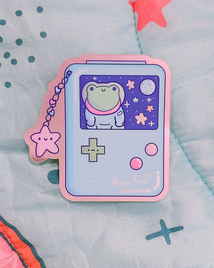 A kawaii matte mirror with orange chrome border vinyl sticker featuring a blue game boy style gaming handheld console with pink buttons and an unhappy green cross hair button. The image also has a happy pink star charm with three hearts. The screen features a frog astronaut in space with stars and moon. An original Prism Pear Designs design.