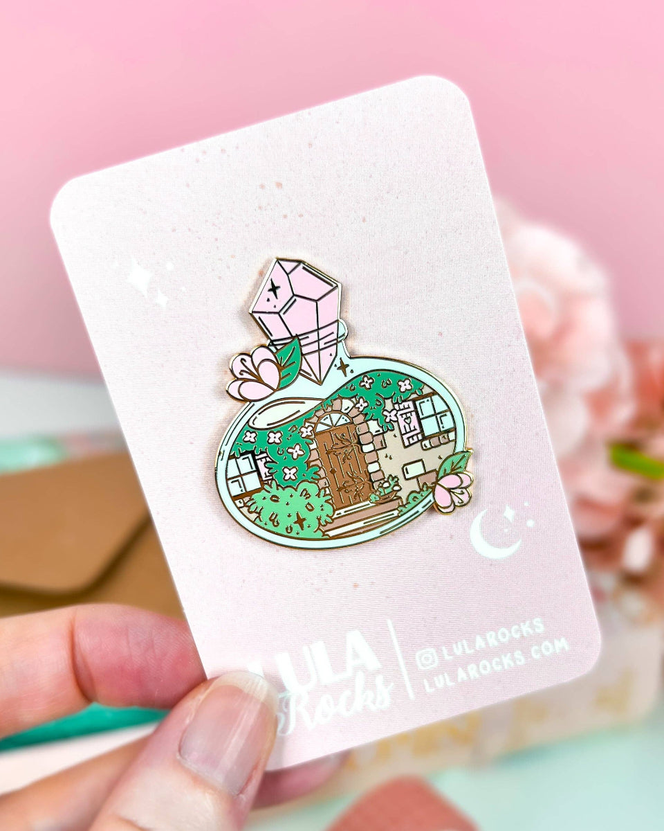 A cottage door with plants, vines, windows, and stairs inside a potion bottle.  The potion bottle is decorated with flowers and closed with a pink gem.  The design is on a hard enamel pin with gold plating.  The pin is on a pink and white lula rocks backing card.