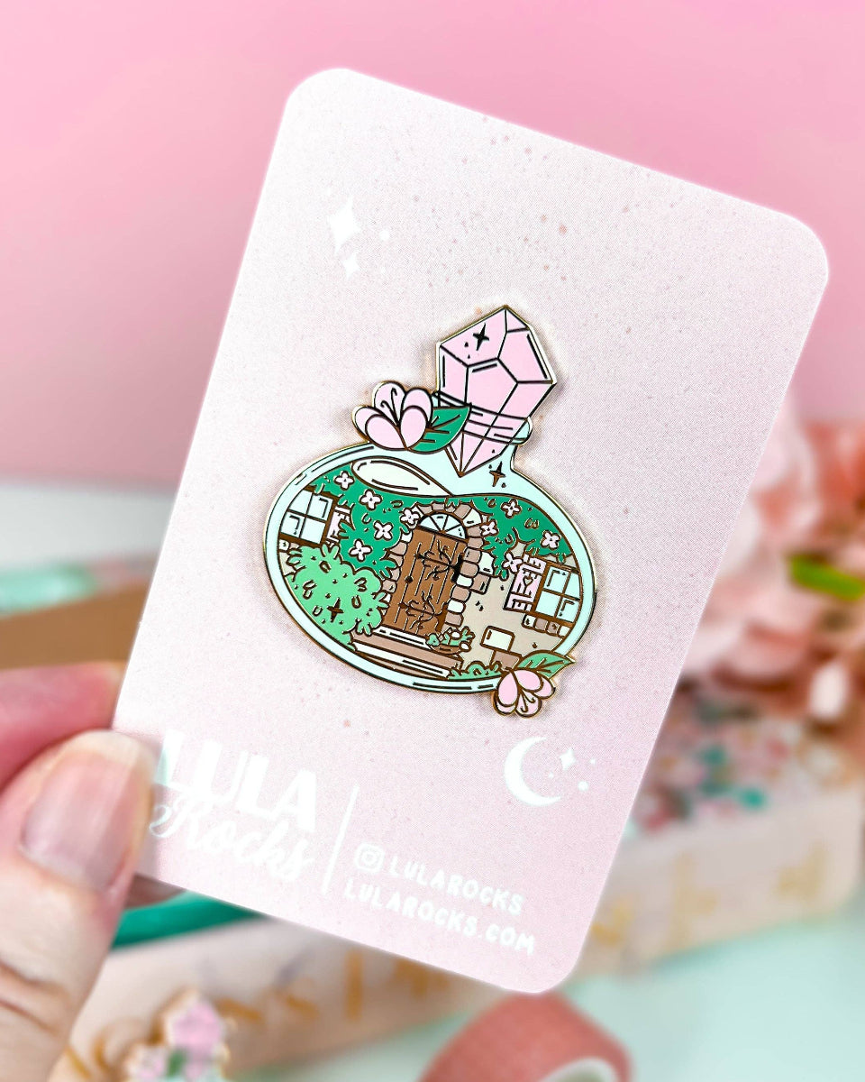 A cottage door with plants, vines, windows, and stairs inside a potion bottle.  The potion bottle is decorated with flowers and closed with a pink gem.  The design is on a hard enamel pin with gold plating.  The pin is on a pink and white lula rocks backing card.