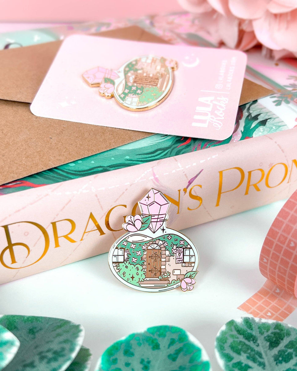 A cottage door with plants, vines, windows, and stairs inside a potion bottle.  The potion bottle is decorated with flowers and closed with a pink gem.  The design is on a hard enamel pin with gold plating.  The pin is leaning backwards on the spine of a pink and gold foiled book.  On top of the book is another pin on a pink and white backing card.