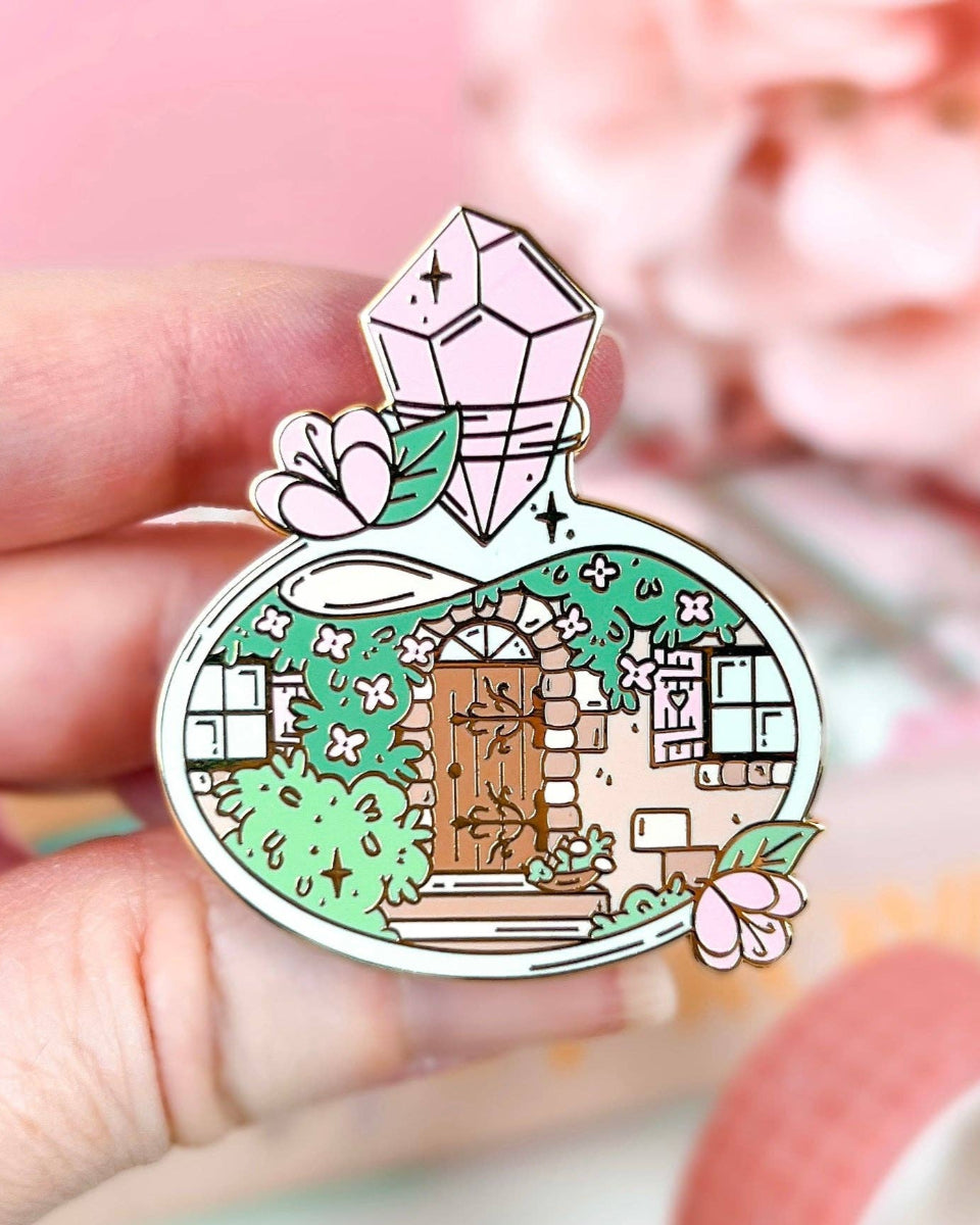A cottage door with plants, vines, windows, and stairs inside a potion bottle.  The potion bottle is decorated with flowers and closed with a pink gem.  The design is on a hard enamel pin with gold plating.  The pin is being held between 3 fingers.