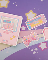 Photo shows a full pack of the vinyl stickers inside the pastel pink game boy style packaging. On the right, there is a pastel pink, blue, and yellow nintendo switch that reads "game over", a pastel pink, blue, yellow, and purple playstation style gaming controller. Both stickers are vinyl.