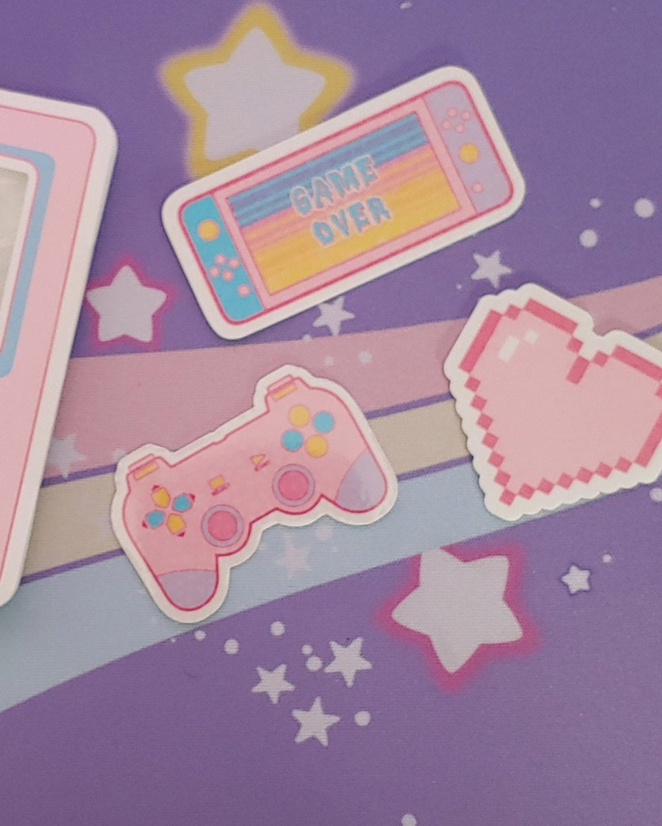 There is a pastel pink, blue, and yellow nintendo switch that reads "game over", a pastel pink, blue, yellow, and purple playstation style gaming controller. There is also a pastel pink pixel heart. All three stickers are vinyl.