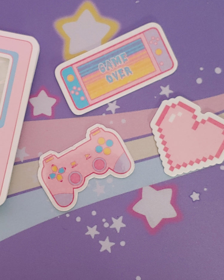 There is a pastel pink, blue, and yellow nintendo switch that reads "game over", a pastel pink, blue, yellow, and purple playstation style gaming controller. There is also a pastel pink pixel heart. All three stickers are vinyl.