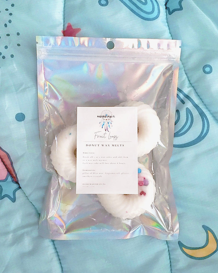 Three donut shaped white swirl with sprinkles wax melts that smell like fruit loops in a holographic resealable pouch that is sitting on a blue, pink, and yellow space blanket.