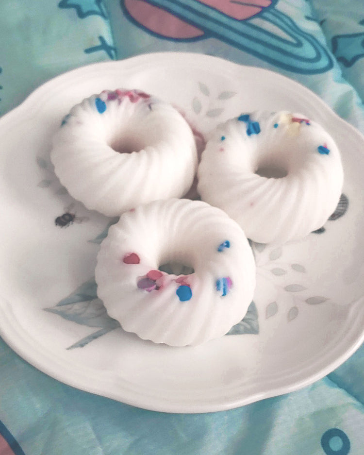 Side view of: Three donut shaped white swirl with sprinkles wax melts that smell like fruit loops on a white and floral plate which sits on a blue and pink space blanket.