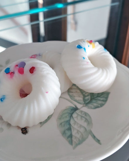 A closer shot of: Three donut shaped white swirl with sprinkles wax melts that smell like fruit loops stacked on a white and floral plate which sits in a glass cupboard.