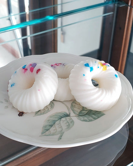 Three donut shaped white swirl with sprinkles wax melts that smell like fruit loops stacked on a white and floral plate which sits in a glass cupboard.