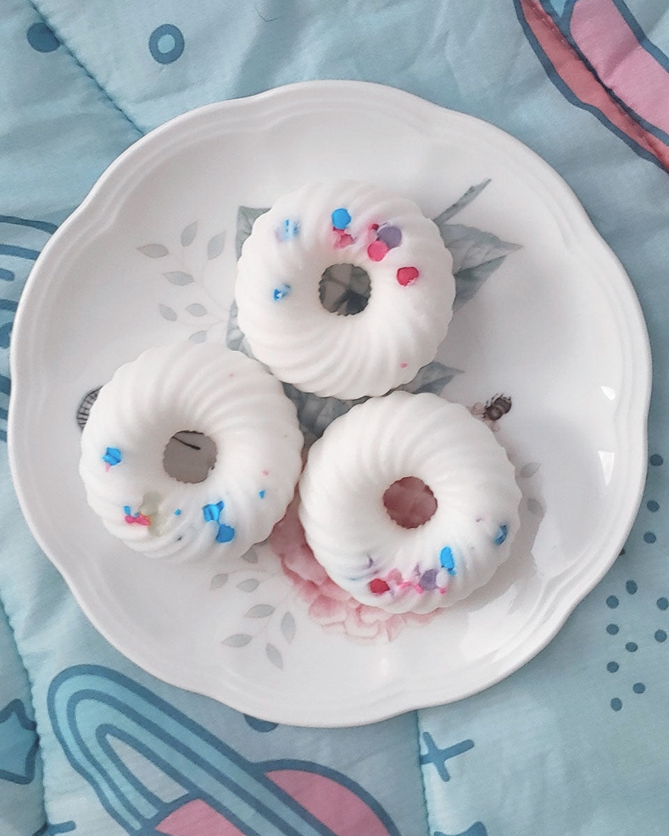 Above lay flat view of: Three donut shaped white swirl with sprinkles wax melts that smell like fruit loops on a white and floral plate which sits on a blue and pink space blanket.
