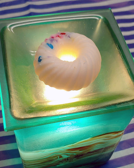 A donut shaped white swirl with sprinkles wax melt that smell like fruit loops sits on top of a green glass wax warmer.  The device is sitting on top of blue and white striped surface.