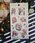 Purple and pink natural magic girls pink foiled sticker sheet sitting on top of a bookmark featuring a candle lit library.  In the corner is a set of yellow fairy earrings.  Surrounded by confetti and gemstones all on top of blue and white striped tissue paper.  Sticker sheet mainly in frame.