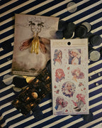Purple and pink natural magic girls pink foiled sticker sheet sitting on top of a bookmark featuring a candle lit library.  In the corner is a set of yellow fairy earrings.  Surrounded by confetti and gemstones all on top of blue and white striped tissue paper.  