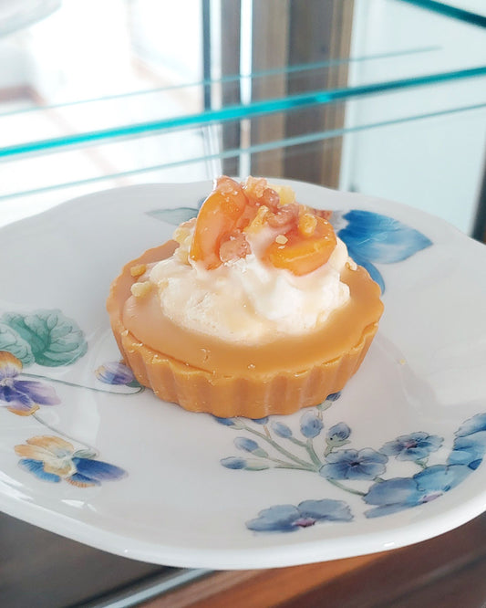 An orange and white max melt appearing to look like a peach pie with orange crust and white whipped cream with beautifully cute peach slices on top.  It's shown on top of a white plate with blue flowers.  Looks like a pie, but is not edible.