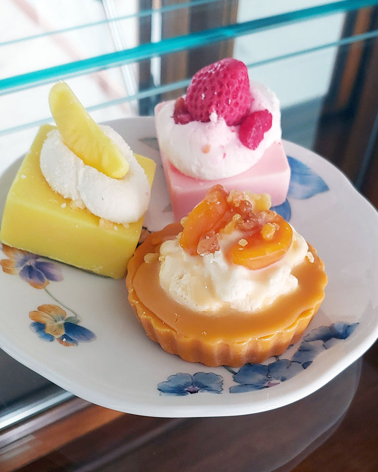 An orange and white max melt appearing to look like a peach pie with orange crust and white whipped cream with beautifully cute peach slices on top. It is shown on a plate with blue flowers next to the lemon bar and strawberry shortcake wax melts. None of the wax melts are edible.