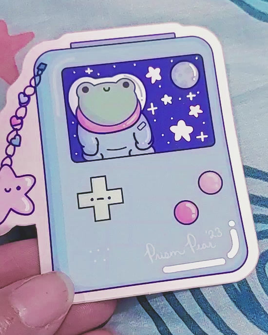 A brief boomerang of a kawaii matte mirror with orange chrome border vinyl sticker featuring a blue game boy style gaming handheld console with pink buttons and an unhappy green cross hair button.  The image also has a happy pink star charm with three hearts.  The screen features a frog astronaut in space with stars and moon. To show reflective and chrome effect on the sticker.