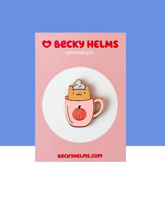 A little kawaii smiling orange cat sitting in a pastel pink coffee mug.  The mug has an orange and black pumpkin printed on it.  The cat has a white glitter whipped topping on it's head. This design is printed on a hard enamel pin with gold-toned finish.