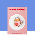 A little kawaii smiling orange cat sitting in a pastel pink coffee mug.  The mug has an orange and black pumpkin printed on it.  The cat has a white glitter whipped topping on it's head. This design is printed on a hard enamel pin with gold-toned finish.