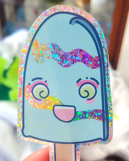 A light blue popsicle sticker with light green swirly eyes, hot pink eye shadow, and light pink rosy cheeks.  The popsicle has a huge kawaii grin on its face.  This glossy vinyl glitter sticker has a glitter border, and two rainbow wavy glitter line details across it's body.  It has a beige stick at the bottom.