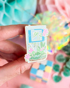 Hand holding a hard enamel pin shaped like a game boy with floral green and pink design against a blurred background
