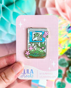 Lula Rocks hard enamel pin shaped like a game boy with pink and green floral on a pink card with a colorful background