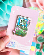 Hard enamel pin shaped like a game boy with pink and green floral design on a pink card held by a hand, with a colorful background.