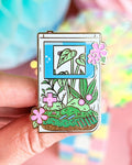 Hard enamel pin shaped like a game boy with plant design held by a hand against a colorful blurred background