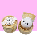 A cute kawaii smiling white bao keychain with rosy cheeks sits inside its open steamer packaging.