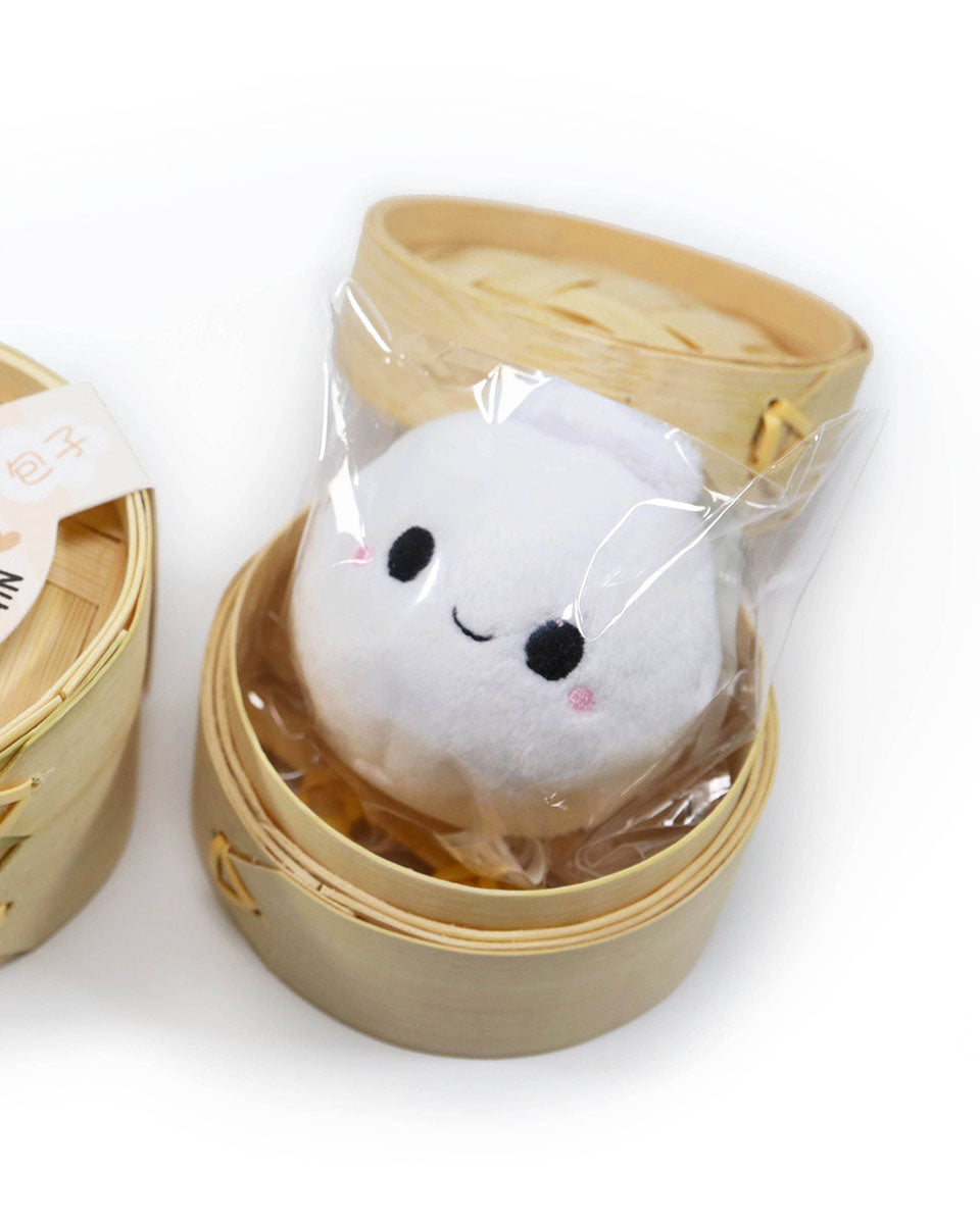 A cute kawaii smiling white bao keychain with rosy cheeks sits inside its open steamer packaging.