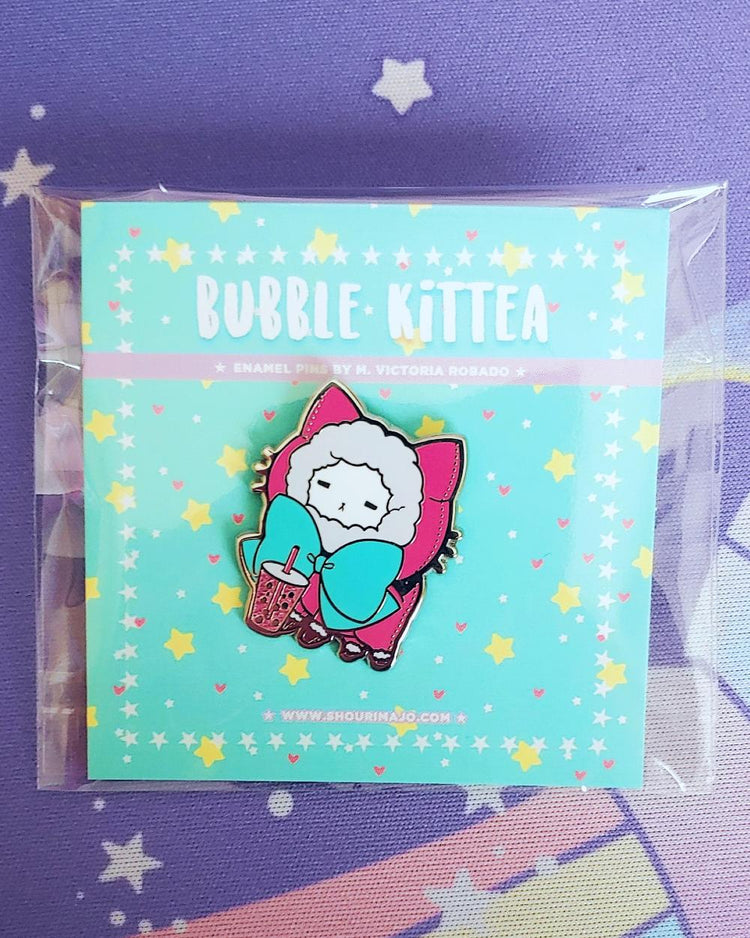 A white kitty cat in a hot pink sweater hoodie pull over with fur lined hood with a teal green giant bow. The kitten is drinking a hot pink boba drink. All of this art is on a had enamel pin and mint green "bubble kittea" backing card.  Wrapped in plastic,