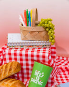 white, pink, orange, green, blue, and yellow gel clicker pens in a picnic basket with green grapes sitting on top of a planner and picnic blanket surrounded by baguettes and a green cup that says "life's a picnic"