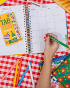 pink, yellow, orange, blue, and white clicker gel pens sitting on a picnic blanket surrounded by zipper bags, sticky notes and a planner.  A woman's hand holding a green pen that reads "you're the cherry on top" about to write something in a planner.