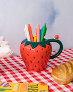 orange, yellow, green, blue, and white click gel pens in a mug shaped like a strawberry on picnic blanket surrounded by flowers and a baguette 