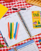 white, orange, green, yellow, blue, and pink gel clicker pens on top of a planner sitting on a picnic blanket surrounded by sunglasses, a baguette, and sticky notes.