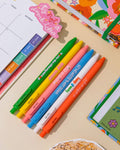 A green click gel pen that reads You're the cherry on top", next to a yellow pen that reads "stay sunny", next to a pink pen that reds "life's a picnic", next to a blue pen that reads "let me write that down", next to a white pen that reads "keep it easy", next to a orange pen that reads "let the delight in" all sitting on a table with different planners are them.