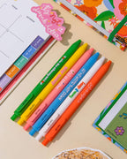 A green click gel pen that reads You're the cherry on top", next to a yellow pen that reads "stay sunny", next to a pink pen that reds "life's a picnic", next to a blue pen that reads "let me write that down", next to a white pen that reads "keep it easy", next to a orange pen that reads "let the delight in" all sitting on a table with different planners are them.