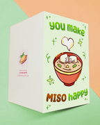 A light green card with a bowl of soup.  Inside the soup are 2 tofu pieces holding hands.  Each has a smile on it's face.  There is a white heart above them.  the card reads you make miso happy in green and brown.  the card also has squiggly lines on it.  You can see the back and it has a cute pie drawing on it.