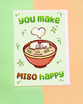 A light green card with a bowl of soup.  Inside the soup are 2 tofu pieces holding hands.  Each has a smile on it's face.  There is a white heart above them.  the card reads you make miso happy in green and brown.  the card also has squiggly lines on it.