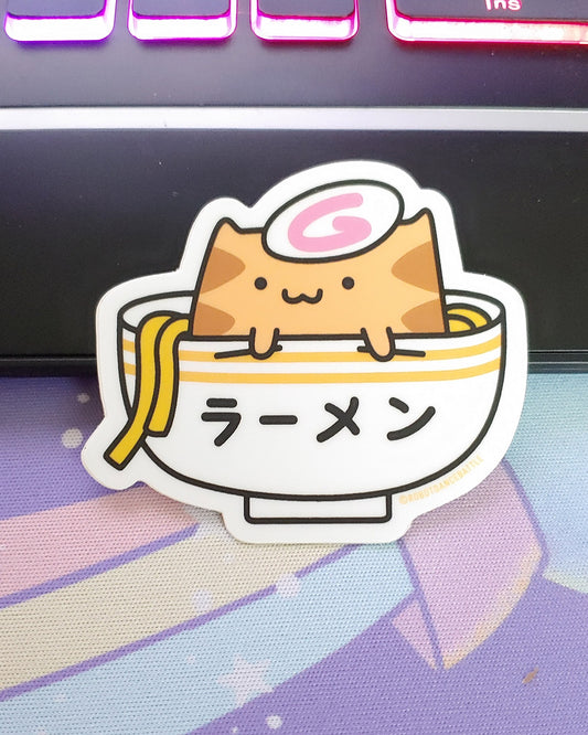 A kawaii orange cat sitting in a striped bowl of ramen noodles. Vinyl sticker.  Designed by small business Robot Dance Battle.