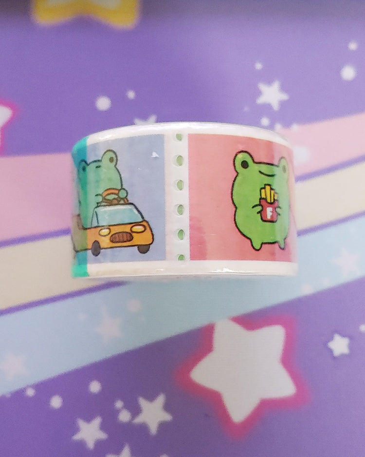 A side of the Froggy Frogs Paper Washi Tape featuring two styles of frogs.  One frog is driving a car and the other is holding some french fries.  The tape is pastel lightly colored stamp washi.  Designed by Robot Dance Battle
